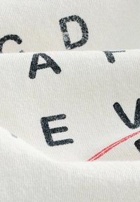 Weißer Stoff mit einer weichen Textur, auf dem schwarze Buchstaben und ein roter Akzent gedruckt sind, angeordnet in einer leicht zerknitterten, geschichteten Präsentation.