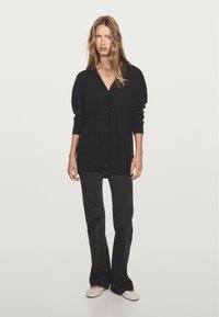 Black cardigan with a V-neck, front buttons, and long sleeves, paired with black flared pants and light-colored shoes. Smooth fabric texture.