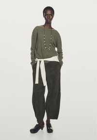 Olivgrön tröja med långa ärmar, vit knytning i midjan, mörkbruna lösa byxor och svarta träskor. Pärlhalsband med jordnära färger.