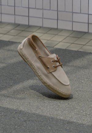 Chaussure bateau décontractée beige et marron avec semelle en corde suspendue au-dessus d'un pavement en asphalte et d'un mur carrelé beige en arrière-plan.