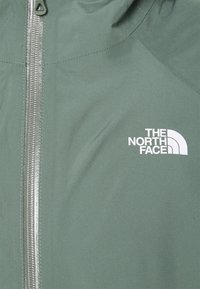 Zielona, wodoodporna kurtka z przednim zamkiem, z białym logo na prawej piersi i gładką, matową teksturą.