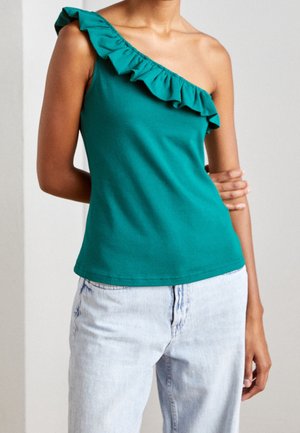 Vrouw die een teal één-schoudertop met ruches draagt en lichtgewassen jeans, staand met een arm over haar borst gekruist en de andere armen langs haar zij.