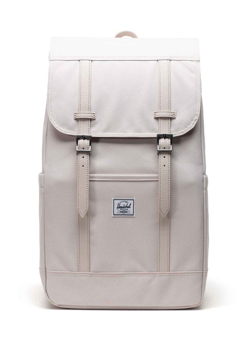 Sac à dos beige avec des sangles à double boucle, rabat supérieur et patch étiquette à l'avant. Présente une forme rectangulaire structurée et une texture de tissu durable.