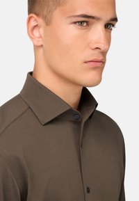 Brūns krekls ar pogām, kuram ir strukturēts apkakles dizains, gluda tekstūra un tumšas pogas. Apģērbs parādīts no sāniem ar tuvplānu.