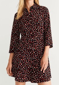 Robe noire présentant un motif abstrait multicolore de carrés rouges, blancs et turquoise. Elle possède un col chemisier et des manches trois-quarts.