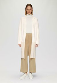 Langer Creme-Cardigan mit offener Vorderseite, kombiniert mit einem weißen Rollkragenpullover und beigen weit geschnittenen Hosen. Weiße Sneakers vervollständigen das Outfit.