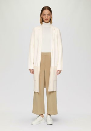 Lange crèmekleurige cardigan met open voorkant, gecombineerd met een witte coltrui en beige wijduitlopende broek. Witte sneakers maken de outfit compleet.