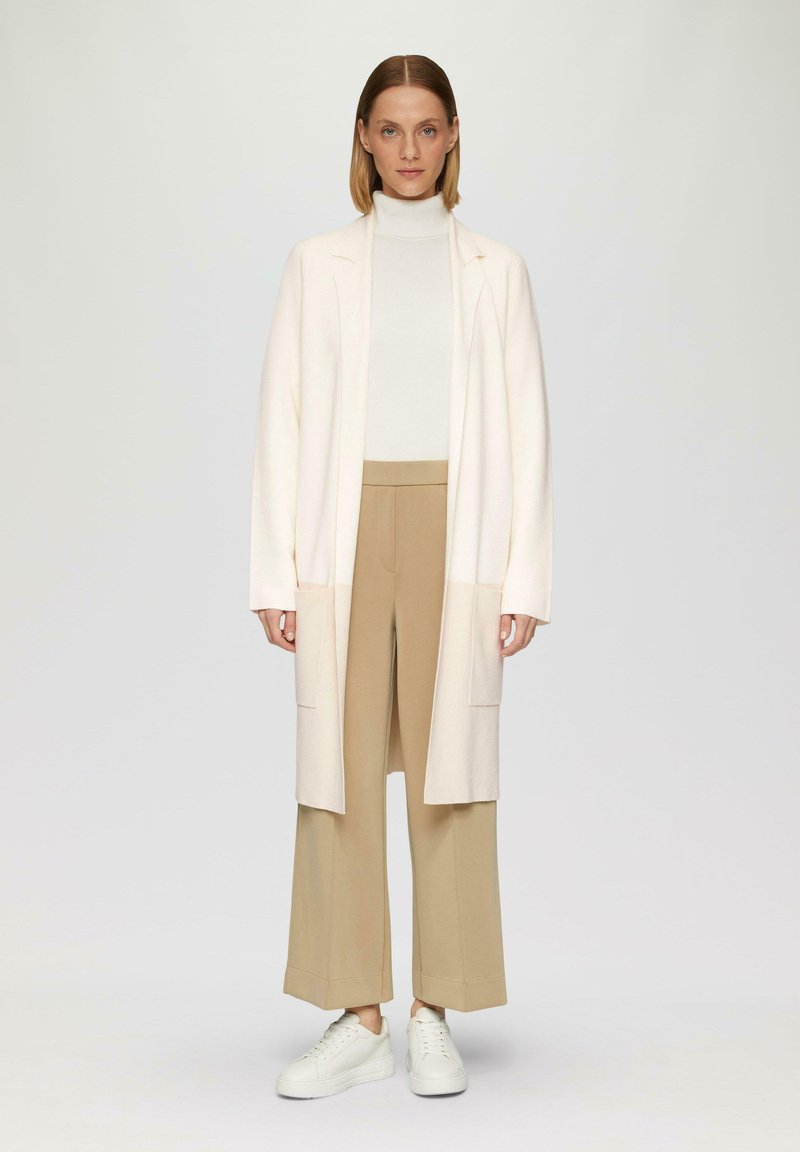 Langer Creme-Cardigan mit offener Vorderseite, kombiniert mit einem weißen Rollkragenpullover und beigen weit geschnittenen Hosen. Weiße Sneakers vervollständigen das Outfit.