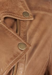 Giacca di pelle marrone con hardware in ottone, caratterizzata da una pattina frontale diagonale e una cerniera. Superficie testurizzata con dettagli di cucitura visibili.