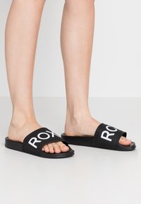 Mustad musta värvi libistasandlid, mis on valmistatud kummist ja millel on lai rihm, millel on valge "ROXY" tekst. Avatud nina disain ja lame talla, sobivad igapäevaseks kandmiseks.