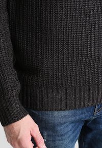 Homme portant un pull en maille côtelée gris foncé et un jean bleu, main détendue le long du corps sur un fond uni.