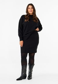 Schwarzes, übergroßes Strickkleid mit langen Ärmeln und rundem Halsausschnitt, kombiniert mit strukturierten schwarzen Cowboy-Stiefeln mit Nietenakzenten.