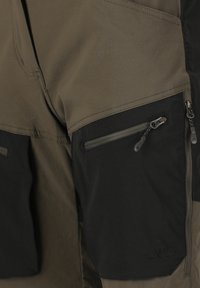 Byxor i olivgrön och svart tyg; har dragkedjefickor och texturerade detaljer, med kontrasterande materialsektioner och en logotyp.