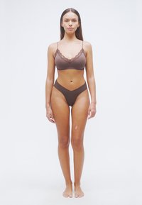 Καφέ bralette με δαντέλα και μπικίνι ψηλόμεσο, φτιαγμένα από απαλό ύφασμα. Το μοντέλο στέκεται ξυπόλυτο σε απλό, φωτεινό φόντο.