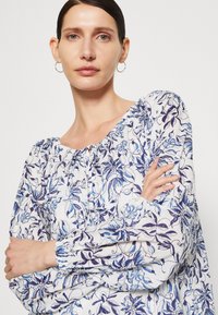 Lett, hvit bluse med blå blomster mønster, samlet halslinning og ballongsleeves med elastiske mansjetter.