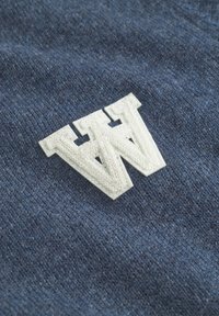 Modrá tkanina s textúrovaným povrchom, na ktorej je v strede biele vyšívané logo "W", prezentujúce vrstvený a trojrozmerný dizajn.