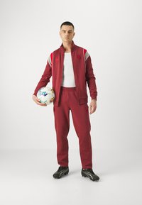 Roter Sportanzug mit einer Jacke zum Reißverschluss und einer elastischen Bundhose, kombiniert mit schwarzen Fußballschuhen und einem mehrfarbigen Fußball.