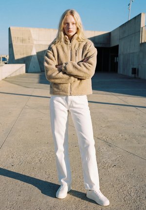 Junge Frau mit langen blonden Haaren steht draußen, trägt eine beige Fleecejacke, weiße Hose und weiße Sneaker, die Arme verschränkt.