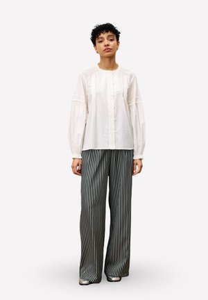 Femme debout, portant une blouse blanche à boutons avec des détails en dentelle et un pantalon large à rayures verticales noir et blanc, des chaussures argentées visibles.