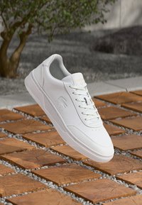 White leather sneaker with a low profile, round toe, lace-up front, and subtle branding on the side. Textured outsole on a tiled background.