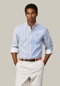 Mladý muž s rukami v vreckách, oblečený v modro-bielej pruhovanej košeli na gombíky, bielych nohaviciach a hnedom opasku.