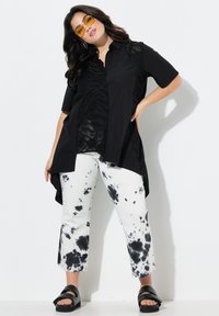 Zwarte shirt met doorschijnende zebra-geïnspireerde textuur, gecombineerd met witte jeans met zwart tie-dye spetters. Model draagt gele zonnebril en zwarte sandalen.