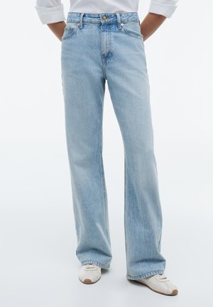 Lichtblauwe jeans met rechte pijpen, gedragen met witte sneakers, handen in de zakken en een gedeeltelijk zichtbaar wit longsleeve-shirt.