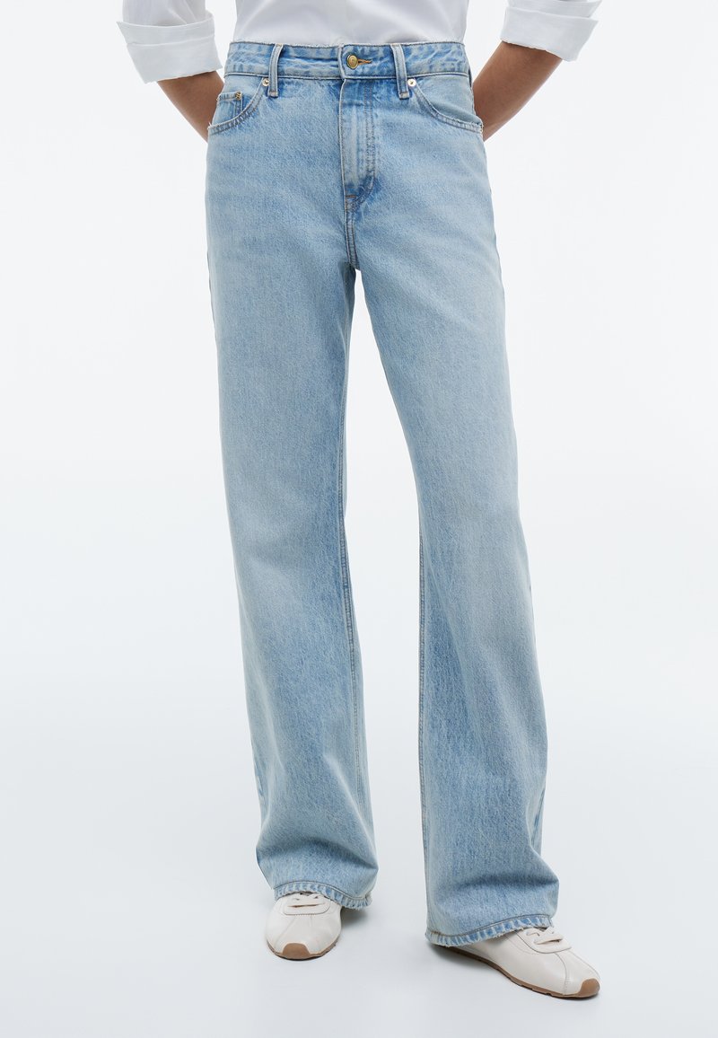 Hellblaue Jeans mit geradem Bein, getragen mit weißen Sneakers, Hände in den Taschen und ein teilweise sichtbares weißes Langarmshirt.