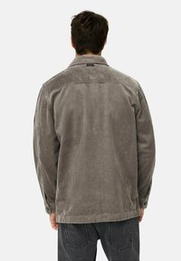 Man in een grijze corduroy jas en zwarte jeans, van de achterkant gezien tegen een effen achtergrond.