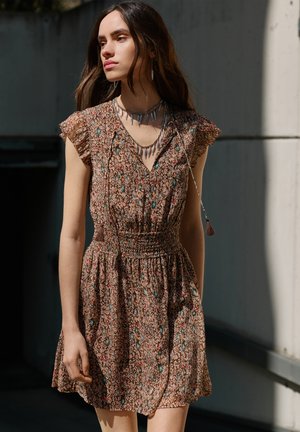 Donna che indossa un vestito corto a maniche corte con vita arricciata e collane sovrapposte a forma di denti, in piedi alla luce naturale contro un muro scuro.