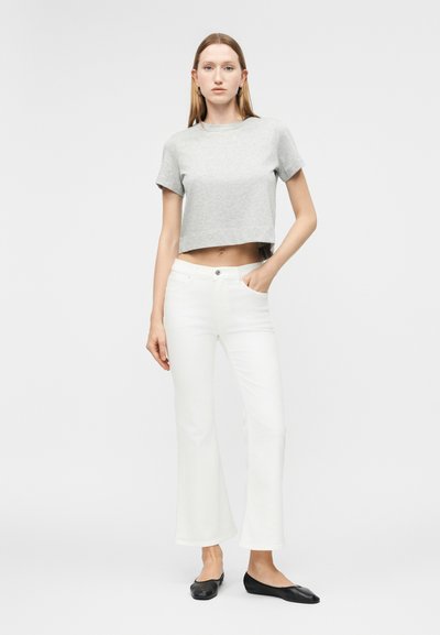 Μια νεαρή γυναίκα στέκεται με το χέρι στην τσέπη, φορώντας γκρι κοντομάνικη μπλούζα, λευκά παντελόνια καμπάνα και μαύρα φλατ παπούτσια, μπροστά σε απλό φωτεινό φόντο.