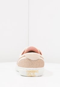 Zapatilla brillante de color rosa dorado con suela blanca, acentos de ante suave y etiqueta de logo. Diseño redondeado con un acabado texturizado en la parte superior.