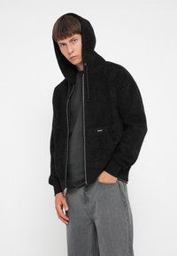 Sort fleece zip-up hoodie med foret hætte, sidelommer og ribbede manchetter. Båret over en mørk skjorte, præsenteret med grå jeans.