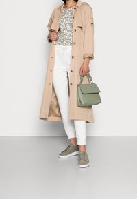 Beige trenchcoat med knapper, grå blomstret top, hvide joggers, grøn håndtaske og grå sneakers med hvide såler. Glatte teksturer og casual design.