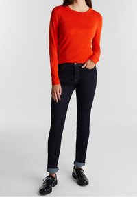 Red long-sleeve sweater with a round neckline, paired with dark blue slim-fit jeans, featuring rolled cuffs and black lace-up shoes.