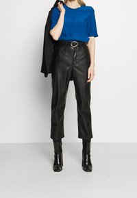 Niebieska bluzka z krótkim rękawem w połączeniu z wysokimi skórzanymi spodniami w kolorze czarnym i czarnymi, eleganckimi botkami na obcasie. Całość uzupełnia czarna marynarka noszona na ramieniu.