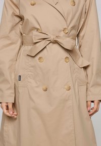 Cappotto beige con design doppiopetto, grandi bottoni e cintura annodata in vita. Presenta tasche laterali e una texture liscia.