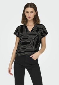 Blusa nera a maniche corte con motivi geometrici bianchi, design a V, e vestibilità rilassata, indossata con pantaloni neri.