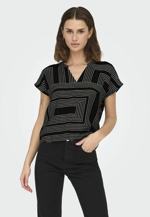 Blusa nera a maniche corte con motivi geometrici bianchi, design a V, e vestibilità rilassata, indossata con pantaloni neri.