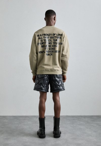 Olivgrüner Pullover mit schwarzem Druck auf dem Rücken, kombiniert mit schwarzen gesteppte Shorts und schwarzen Knöchelschuhen.