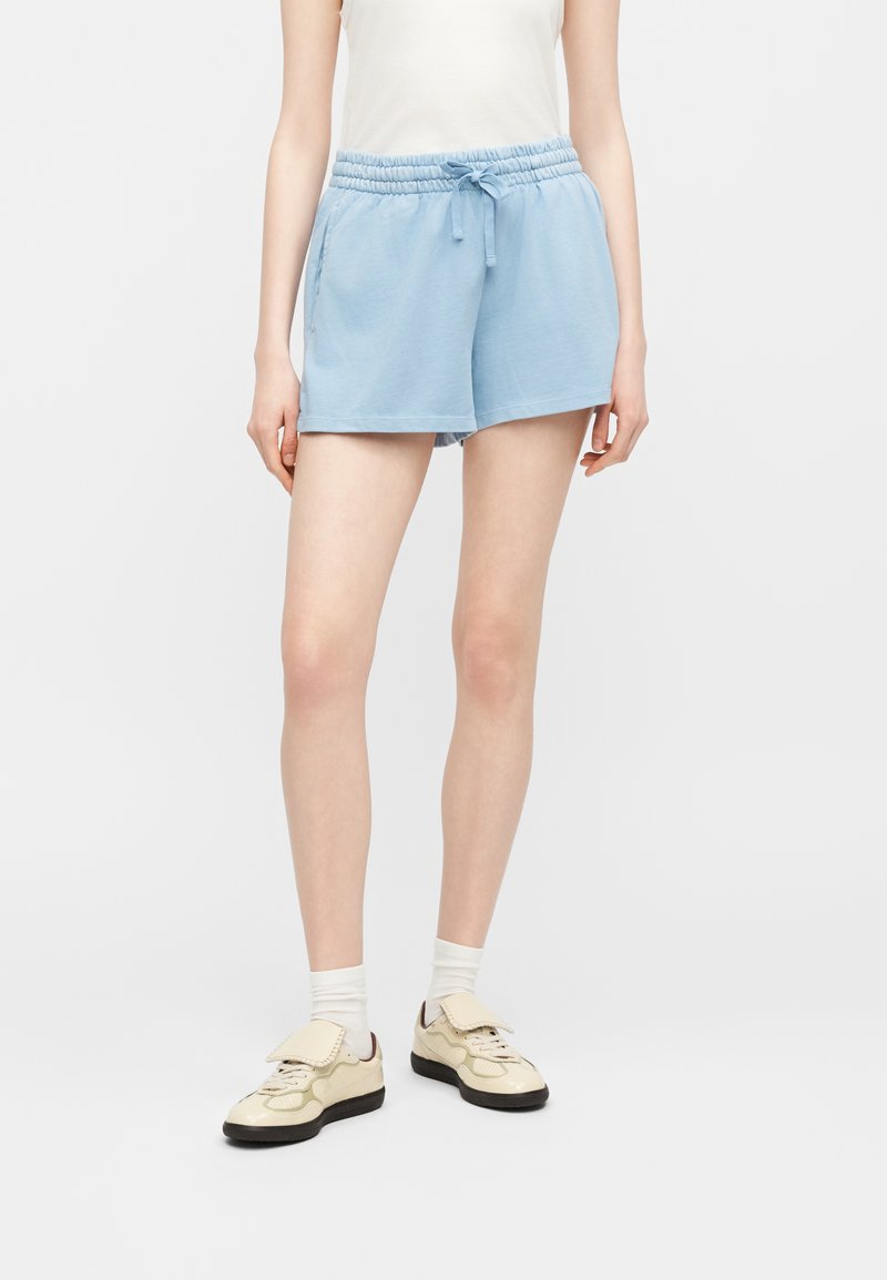 Personne portant un short à cordon de serrage bleu clair, un haut sans manches blanc, des chaussettes blanches et des baskets crème avec des semelles noires, debout sur un fond blanc uni.