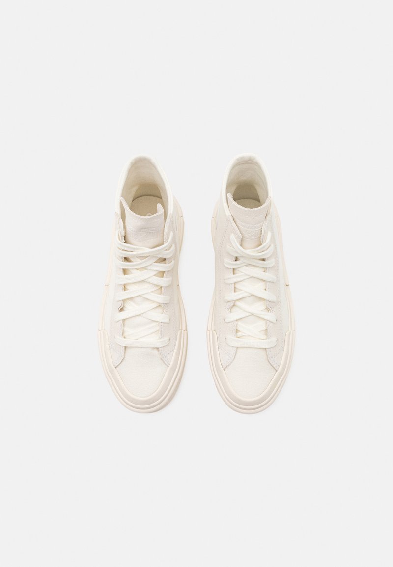 Sneakers high-top in canvas crema con lacci in cotone piatti, suole in gomma testurizzata e collari imbottiti. Design minimalista senza logo visibile.