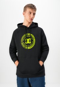 Young man with hands in pockets wearing black hoodie with neon yellow circular DC Shoe Company logo, paired with blue jeans, looking sideways.
