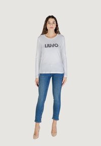 Il modello indossa una maglietta bianca a maniche lunghe aderente con il logo "LIUJO", abbinata a jeans blu e tacchi color pelle, su uno sfondo neutro.