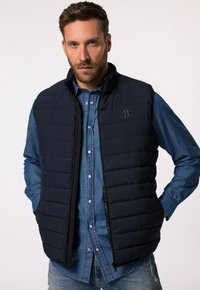Marinefarbener gepolsterter Weste mit hohem Kragen und schwarzem Reißverschluss. Darüber trägt er ein hellblaues Denim-Hemd. Mit dezentem Logodetail auf der Brust.