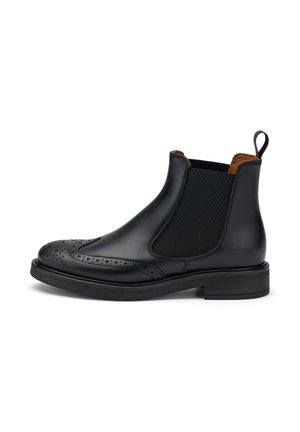 Bota Chelsea de cuero negro con paneles laterales elásticos, detalles de brogue en la puntera y un tacón bajo y apilado. La textura es suave con un acabado mate.