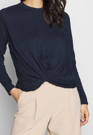 Haut à manches longues bleu marine avec un design en ribbed texturé, présentant un détail avant torsadé. Fabriqué en tissu doux, porté avec un pantalon beige.