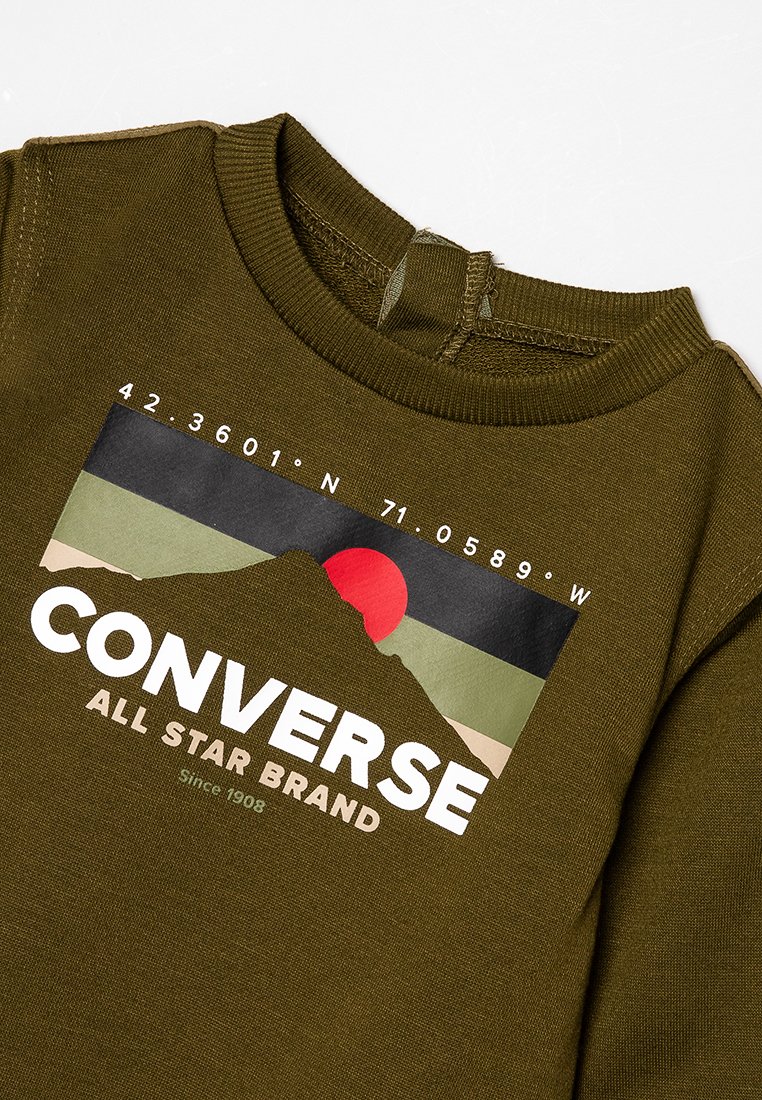 Chemise vert olive avec un col brodé présentant un paysage montagneux, un soleil rouge, des coordonnées et le texte "MARQUE CONVERSE ALL STAR".