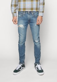 Jeans de denim azul claro con corte ajustado, detalles desgastados con bordes deshilachados y una textura ligeramente descolorida. Llevados con zapatillas negras, blancas y verdes.