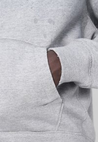 Sweat-shirt gris en tissu doux avec une poche kangourou. Comprend des poignets et un ourlet côtelés, présentant une légère usure et des taches d'eau.